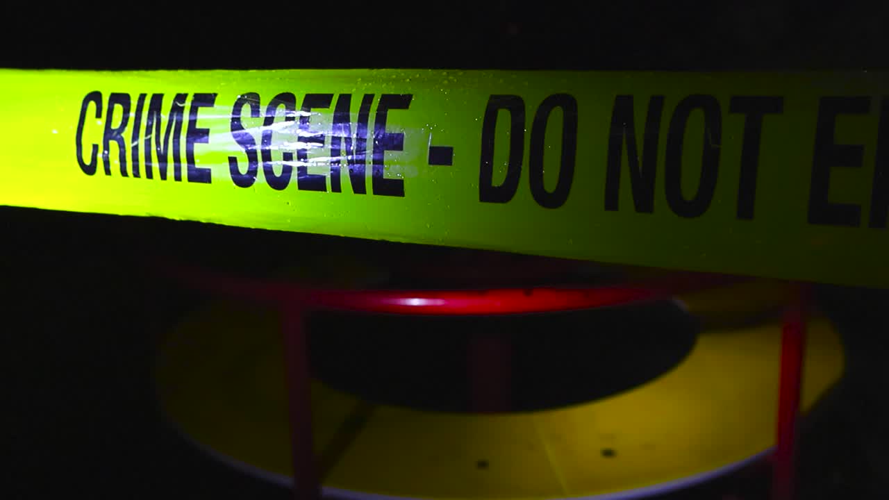Close up or closeup footage of a yellow colored Crime Scene Do Not Enter police caution tape ribbon pulled in front of a merry go round spinning in a dark and rainy night, raindropds on the tape