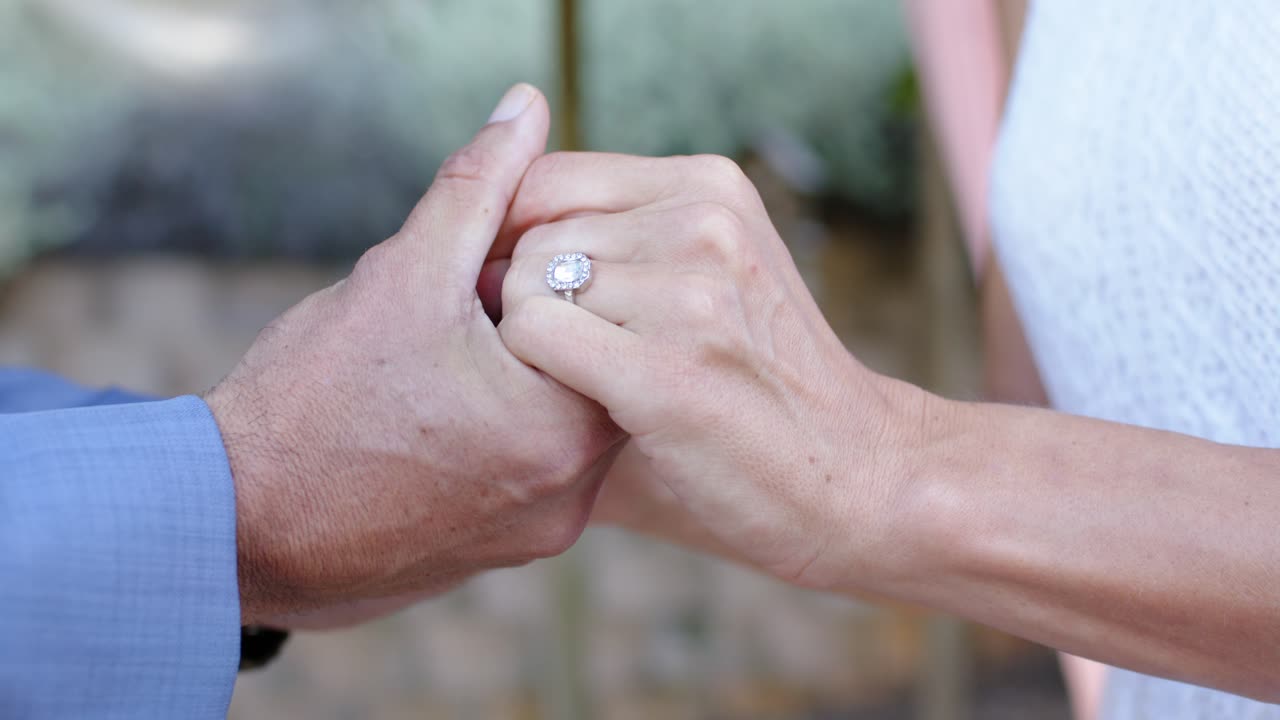 Couple after ring exchange, man in blue shirt steadying woman's hand presenting halo ring outdoors