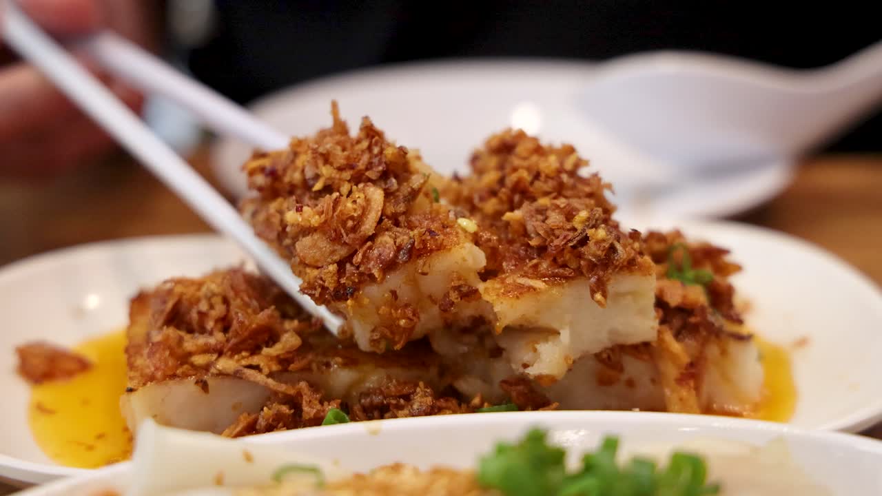 A hand uses chopsticks to lift a piece of fried turnip cake garnished with crispy shallots from a plate in a warmly lit Singapore restaurant