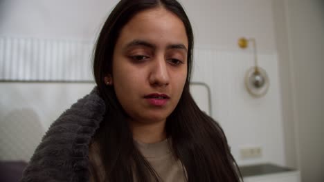 Portrait-of-a-sad-sick-brunette-girl-with-a-cold-in-a-brown-sweater-and-a-dark-gray-blanket-who-coughs-behind-her-hand-and-has-a-runny-nose-at-home-in-a-modern-apartment