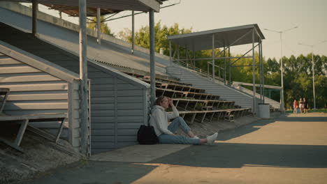 lady seated alone, leaning on stadium pole, looking thoughtful, with empty bleachers beside her, background includes trees, electric poles, and a blurred group of people walking in the distance