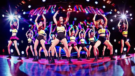 Group of female dancers wearing black and yellow outfits are energetically performing a synchronized dance routine on a stage with a vibrant, colorful backdrop and dynamic lighting