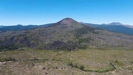 Drone-video-showcases-a-towering-mountain-peak-set-against-a-stunning-blue-sky,-with-additional-hills-across-the-horizon