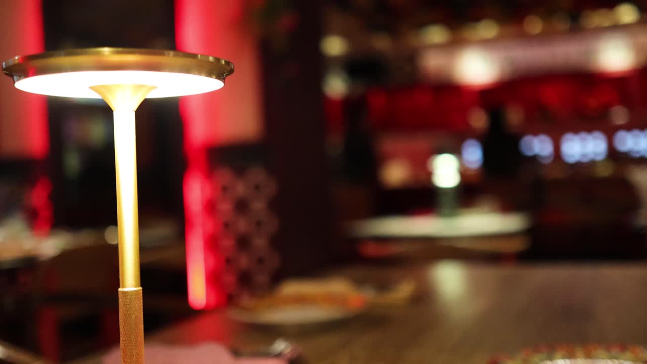 A glowing table lamp stands on a wooden table in a contemporary restaurant, with soft, warm lighting and blurred red-toned background. Static camera, shallow depth of field