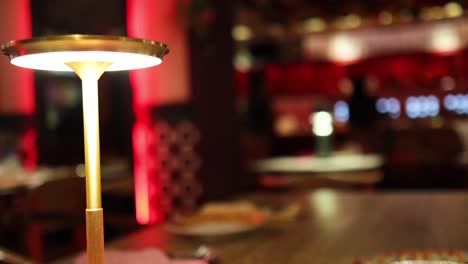 a glowing table lamp stands on a wooden table in a contemporary restaurant, with soft, warm lighting and blurred red-toned background. static camera, shallow depth of field