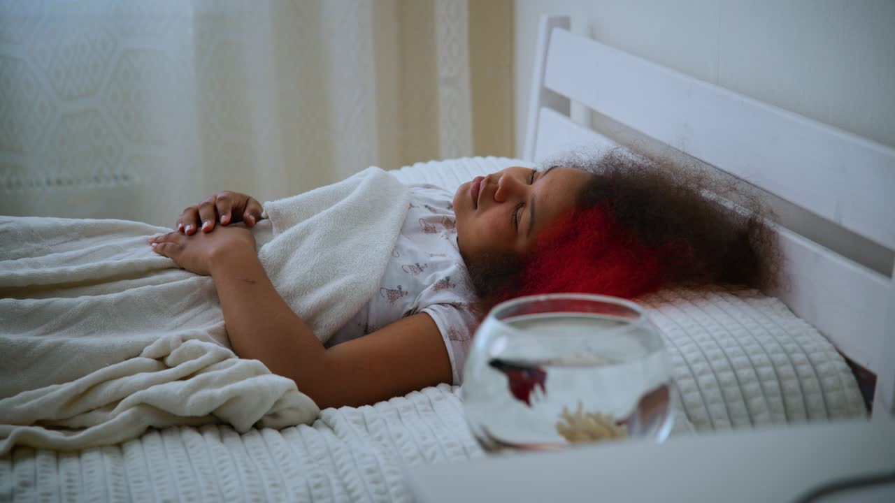 Ung pige med strålende rødt hår, der langsomt vågner i et solbeskinnet soveværelse, strækker armene og nyder et fredfyldt morgenøjeblik nær et akvarium på et pyntet natbord, der indfanger en rolig start på dagen