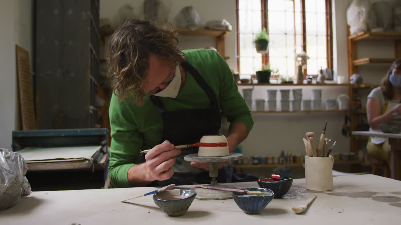 Male caucasian potter wearing face mask and apron using brush to paint pot on potters wheel at potte
