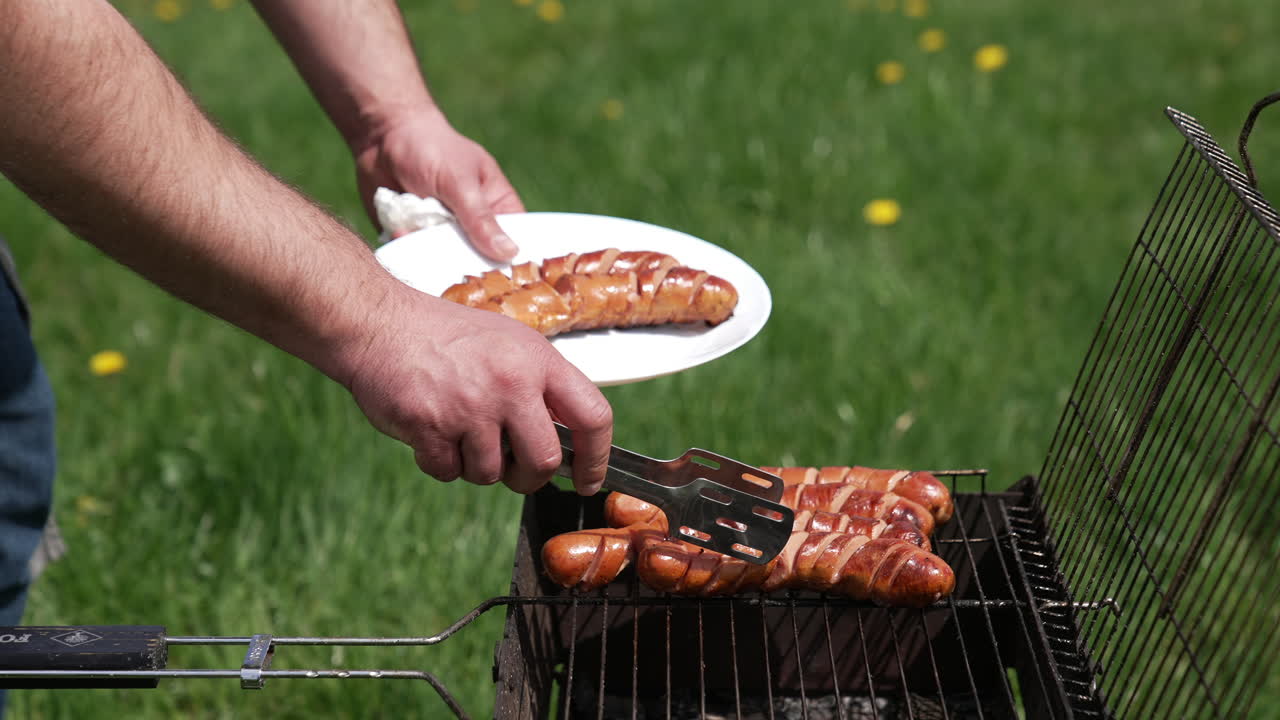 Chef taking off ready sausages from a grill. Man putting delicious roasted hot dog sausages on a plate with a tongs. Barbecue for outdoor lunch.