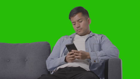 studio shot of young man sitting on sofa scrolling through mobile phone against green screen