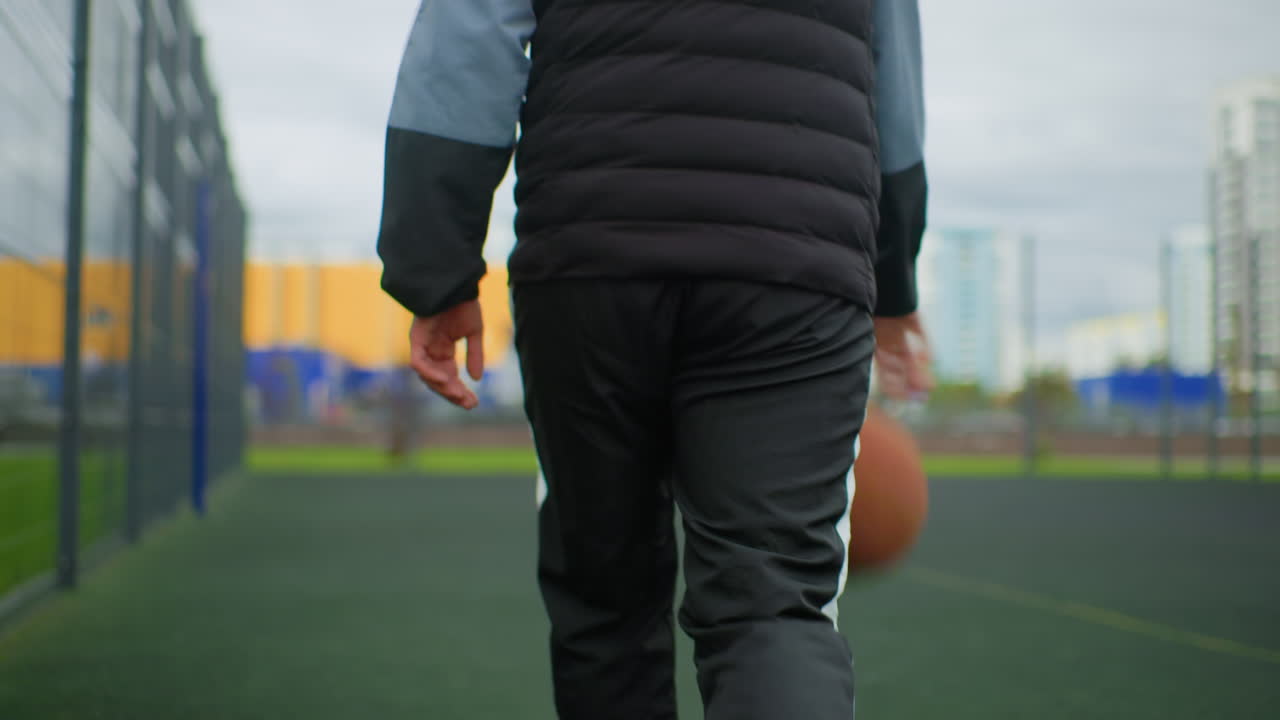 バスケットボールを抱え、中綿ベストとパーカーを着て屋外バスケットボールコートを歩く10代の若者、金網と人工芝がシーンを囲み、曇り空と遠くの都市の建物が都会的な雰囲気を添えています。