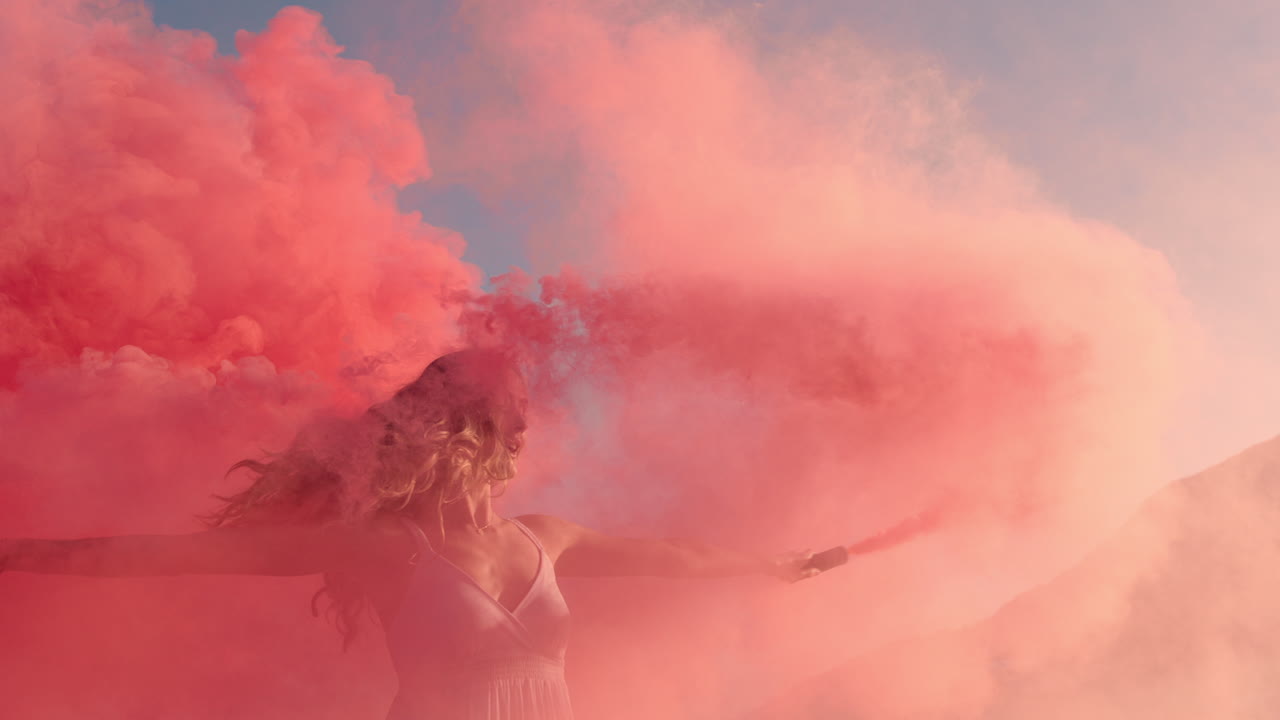 donna che balla con una bomba di fumo rosa sulla spiaggia all'alba celebrando la libertà creativa con una danza giocosa in slow motion