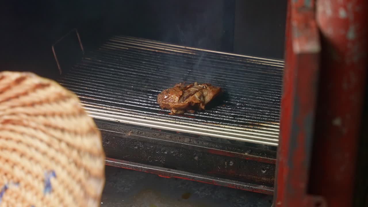 A stationary shot of chicken grilling inside a red metal barbecue smoker, seen over the shoulder of the vendor in a woven straw hat, with smoke rising in Lucban, Quezon Province Philippines