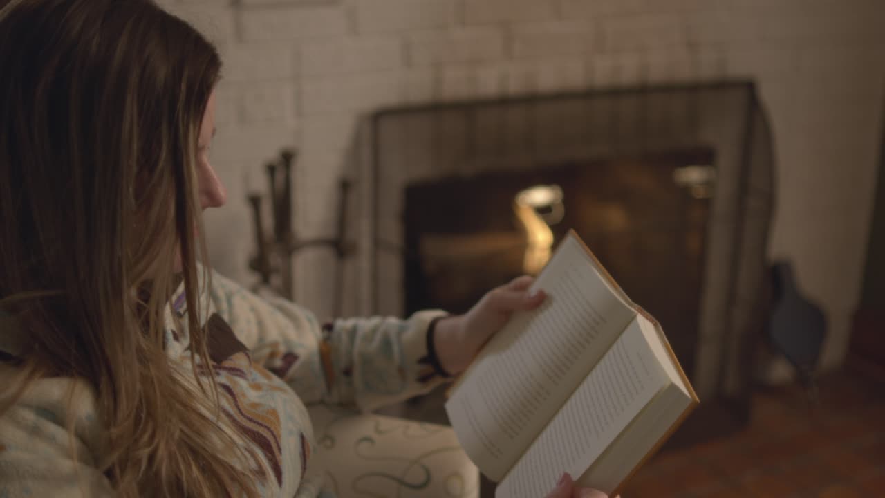 An over the shoulder shot of a woman opening a book and flipping through the pages in front of a fireplace. A warm cozy fire in the background in a fall setting. Shot in 60 frames for slow motion
