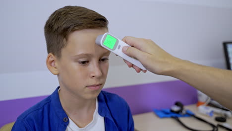 pediatrician or doctor checks boy`s body temperature using infrared forehead thermometer. teenager at doctor`s cabinet. selective focus.
