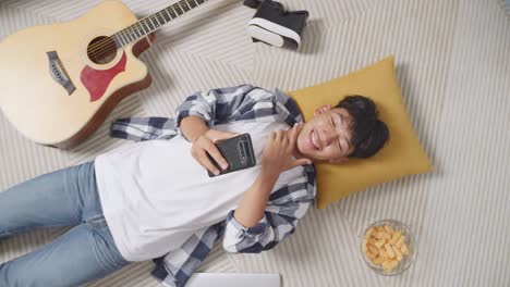 top view of asian teen boy using smartphone taking selfie while lying on carpet on the floor at home. smiling, showing peace and heart gesture