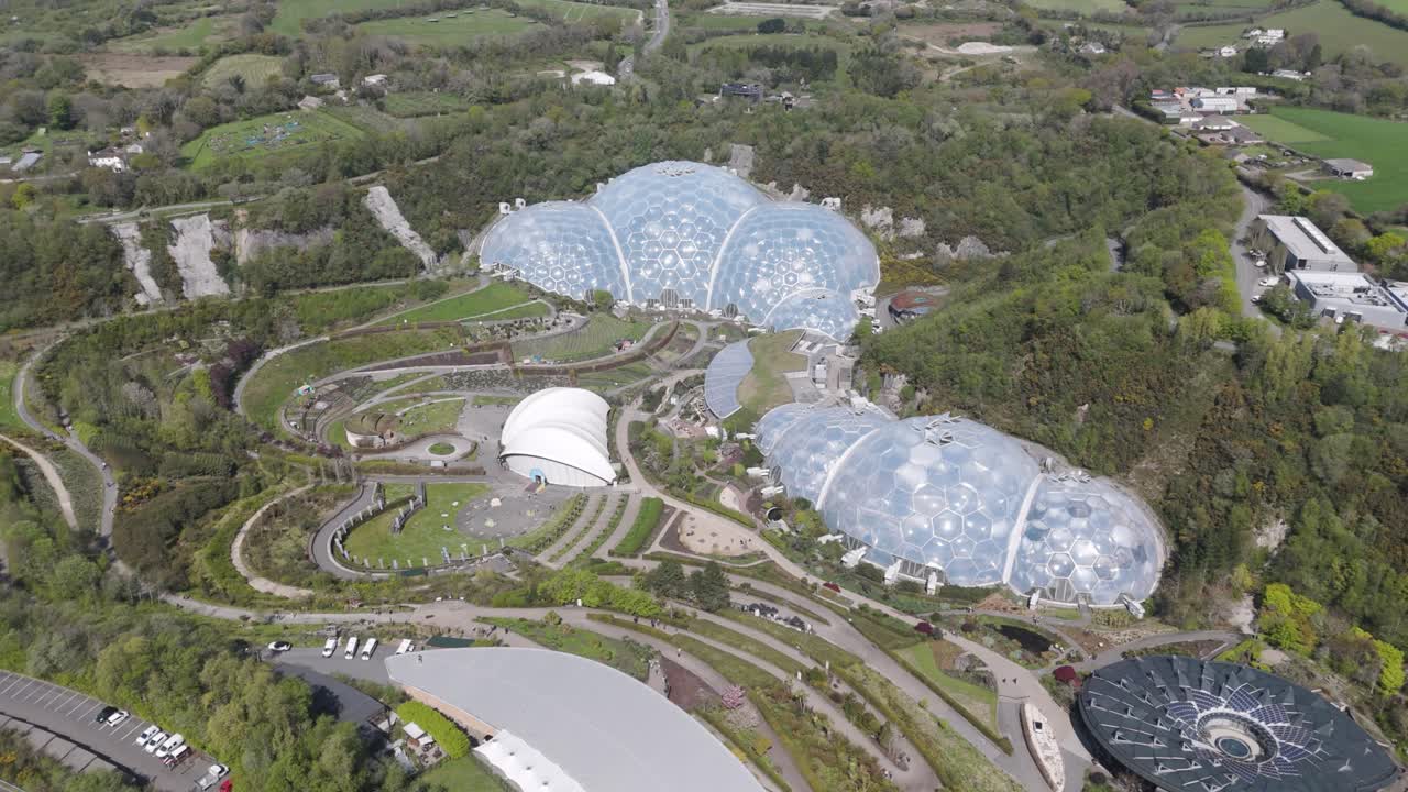 Orbital aerial of Eden Project domes sparkling in sunlight amid spiralling paths and lush plantings inside forested quarry basin, symbolising sustainable environmental education