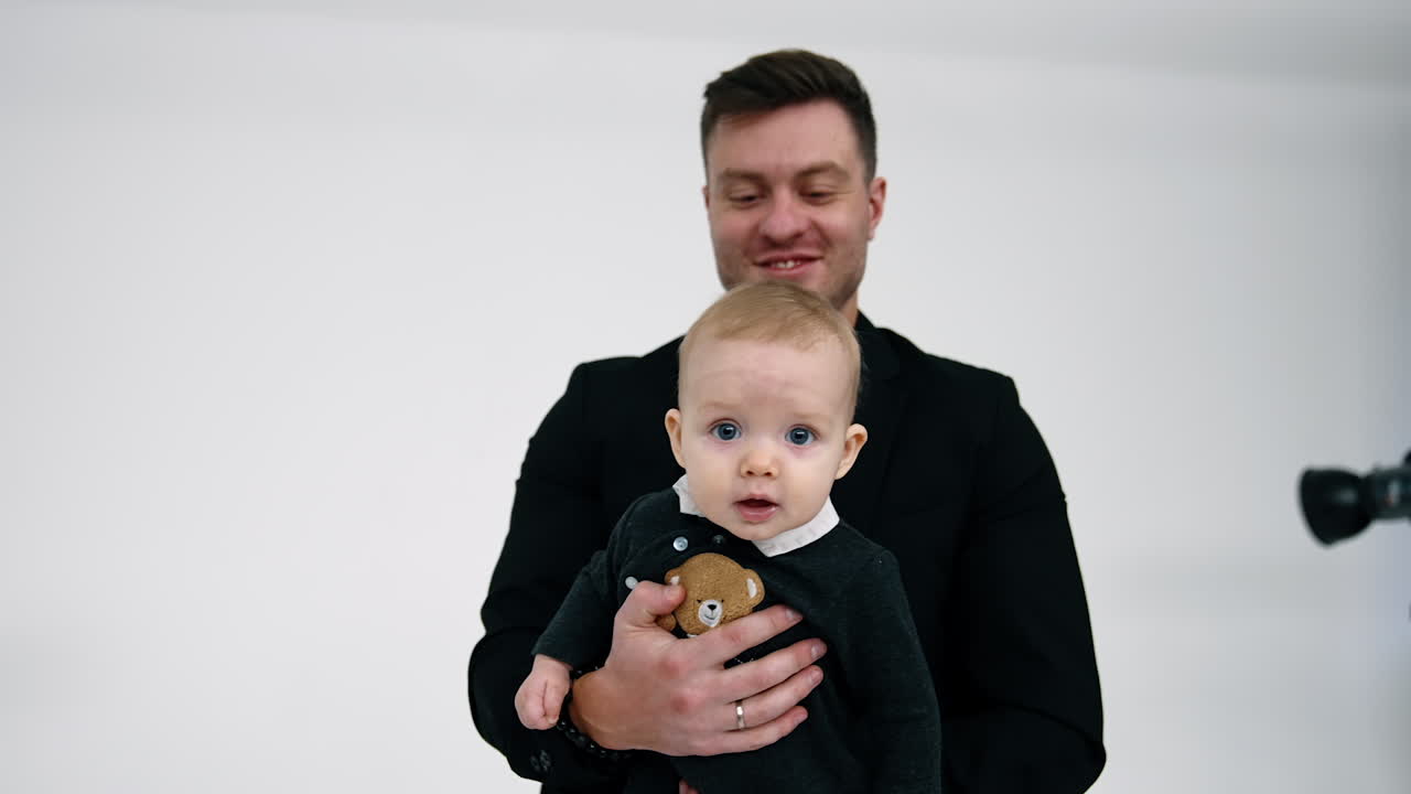 Muscular Caucasian man in black jacket holds a baby in hands. Little child looks around with surprise. White backdrop.
