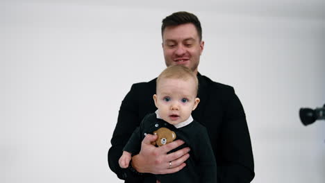 muscular caucasian man in black jacket holds a baby in hands. little child looks around with surprise. white backdrop.