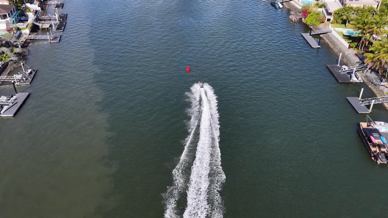 Drone captures jetski racing along residential canal, creating waves, under bright daylight, smooth tracking shot