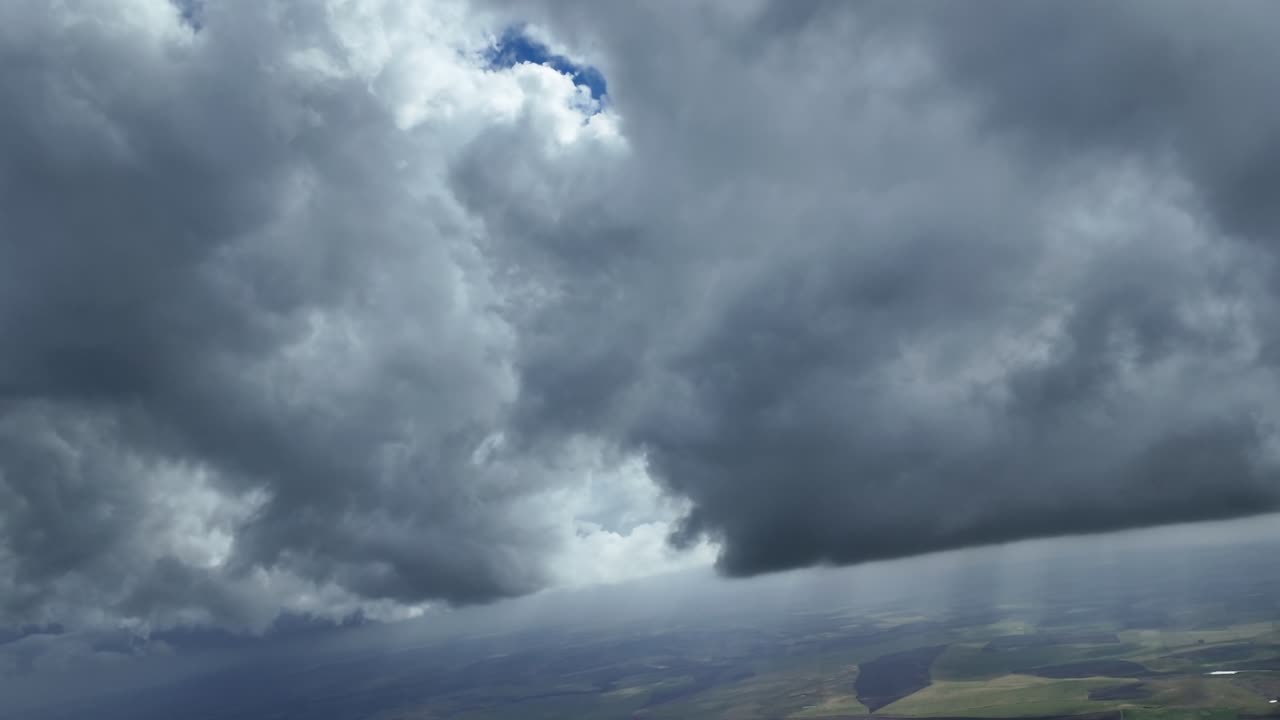 An aerial POV taken from inside an airplane cockpit flying under threatening massive storm clouds with the sunlight piercing through the clods illuminating a rural landscape.