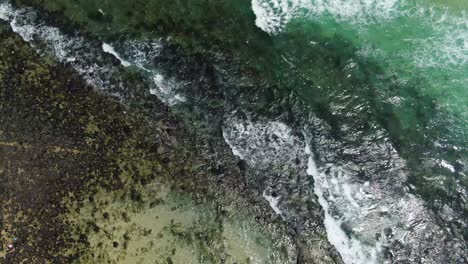 Rising-shot,clear-waters-waves-on-rocky-beach,-bright-day,-surfers-waiting-for-waves