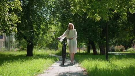 young woman in translucent raincoat strolls with bicycle along sunlit pathway surrounded by greenery, pauses to place stand, remove hood, and lean on handlebars, background features lush trees