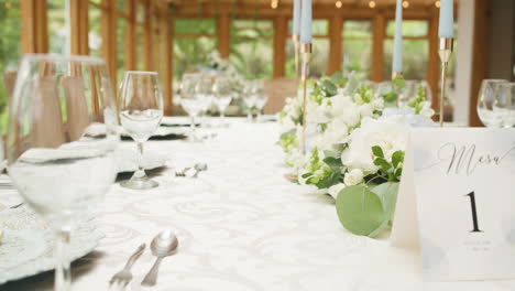 clip of a finely decorated wedding table with a white tablecloth, glass stemware, shiny cutlery, plates, and a delicate floral arrangement in the center.
