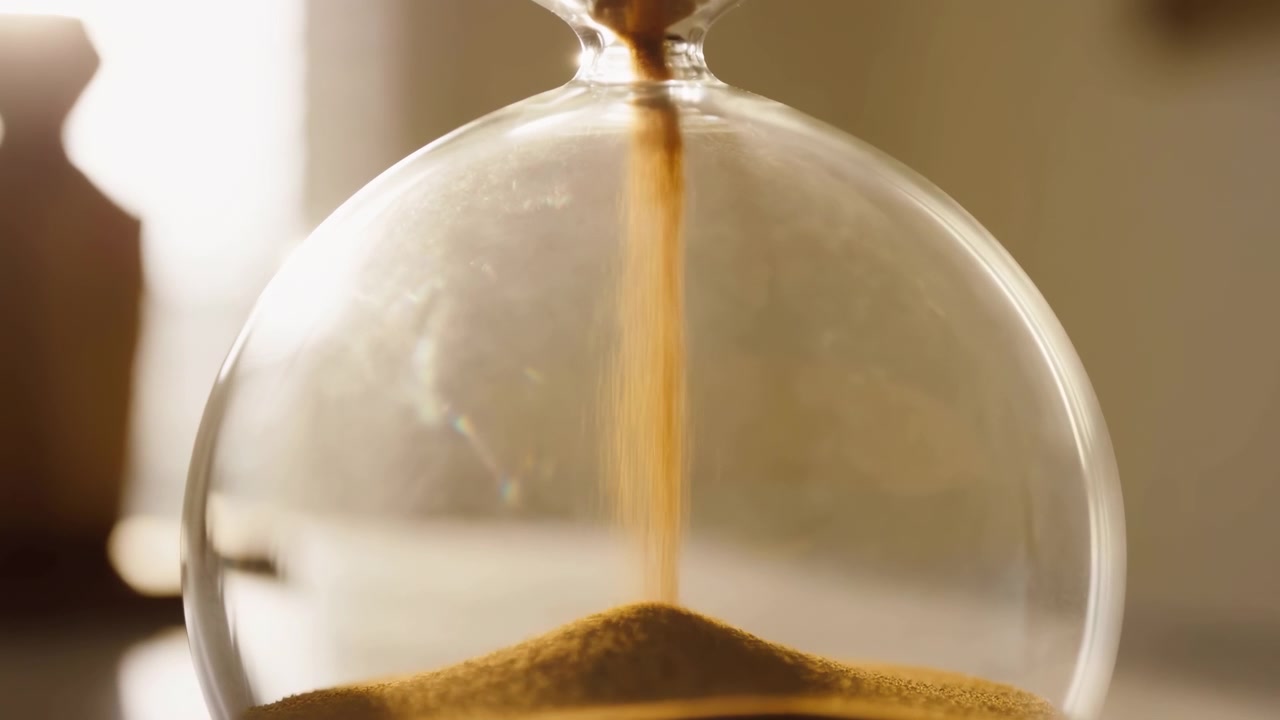 Close-up view of a glass hourglass positioned slightly right of center on a tabletop as warm golden sand streams from the upper bulb into a conical pile in