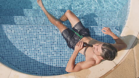 overhead shot of man in swim shorts relaxing in outdoor swimming pool drinking beer