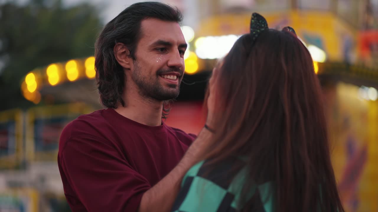 A brunette guy with stubble in a dark red T-shirt and his brunette cosplayer girlfriend on a date in an amusement park that glow brightly. A guy strokes his face and straightens his girlfriend's hair during a date