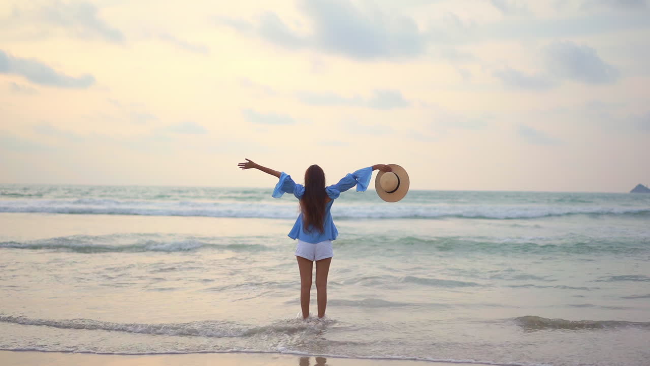 detrás de una mujer solitaria emocionada levantando las manos en la playa de arena frente a las olas del mar y el horizonte, fotograma completo a cámara lenta