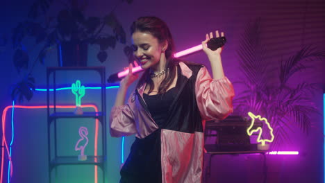 brunette girl dancing cheerfully with led lamp in hands looking at camera on neon wall with colorful lamps