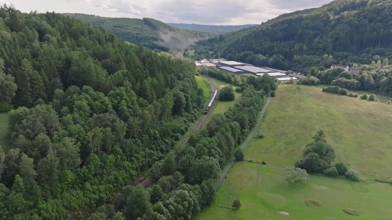 Aerial view of a train traveling through a lush green valley with a factory