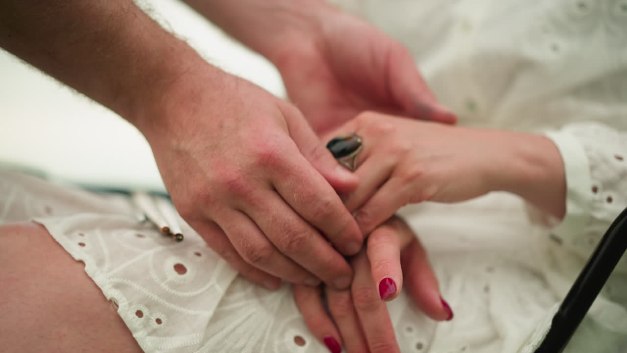 Close-up of a man adjusting a woman's hands, featuring her red nail polish and a distinct ring, set against the backdrop of a white dress