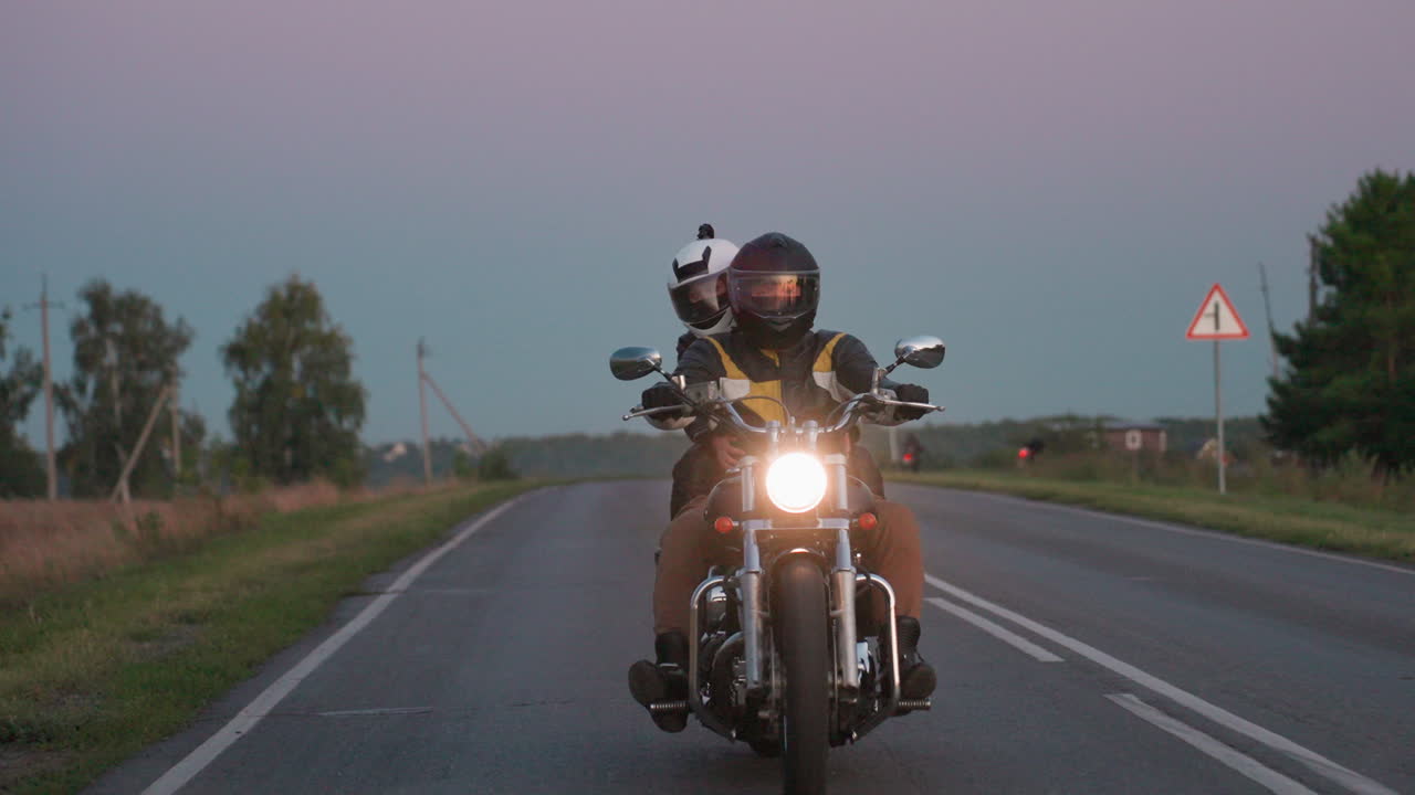 Motorcyclist riding along countryside highway during sunset momentarily greets another rider driving in opposite direction, headlights on, passengers visible, rural background