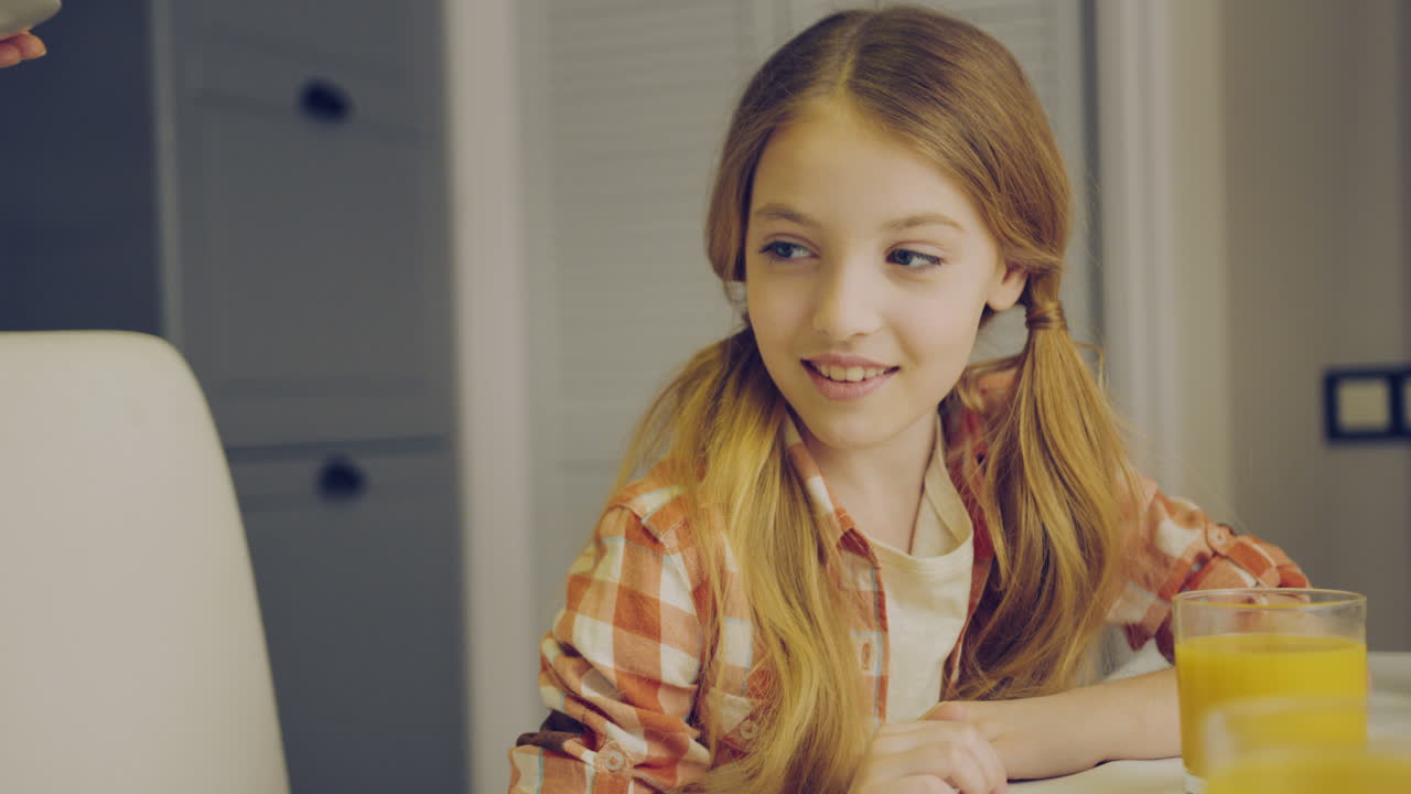 милая блондинка сидит за кухонным столом со стаканом сока, а ее мать приносит ей тарелку кексов. дочь обнимает свою маму. внутри