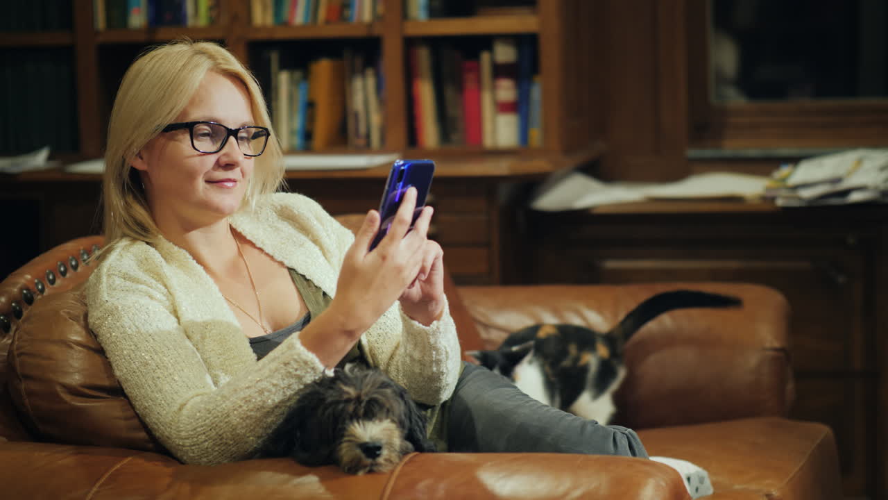 한 여성이 애완동물 옆에 있는 홈 라이브러리에서 스마트폰을 사용합니다.