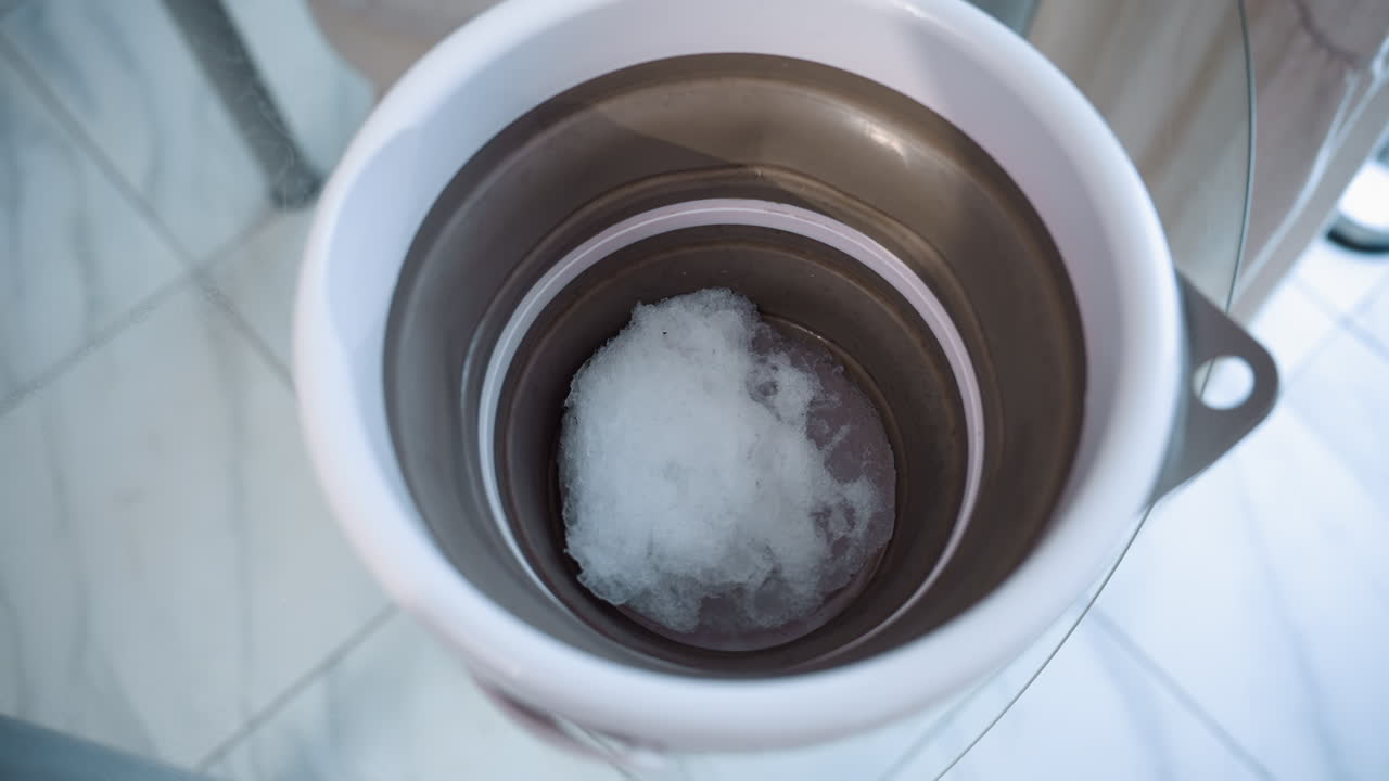 Bucket holding defrosting snow seen from above on plastic topped table under soft light. Blurred setting hints at interior space while delicate slush and moisture glisten inside container