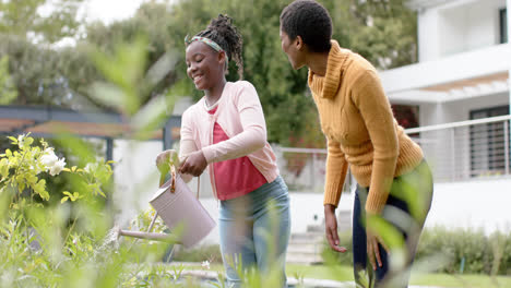 happy african american daughter and mother watering plants in garden, slow motion