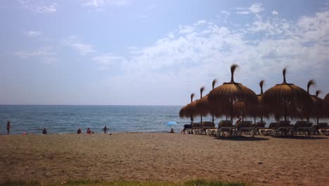 Lapso-De-Tiempo-De-Movimiento-De-La-Playa-De-Marbella-Con-Sombrillas-Tikki-Y-Nubes-Moviéndose-En-Segundo-Plano,-Vacaciones-De-Verano