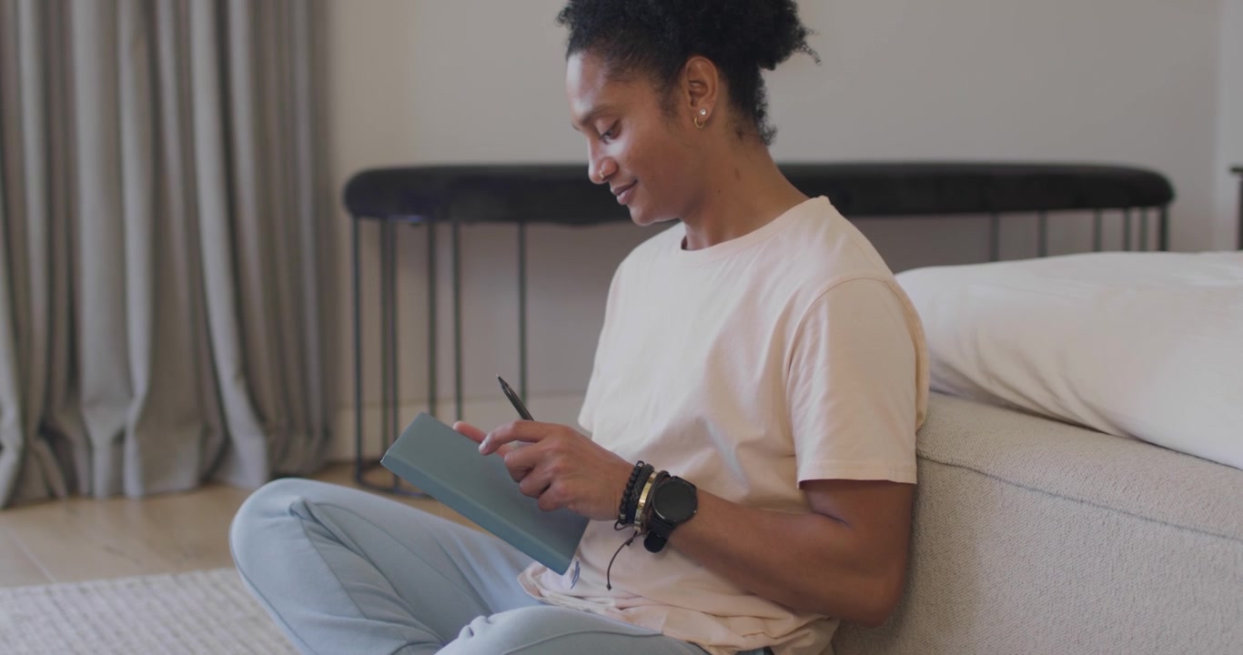 African American woman sitting on floor smiling grabbing teal journal writing with pen for notes