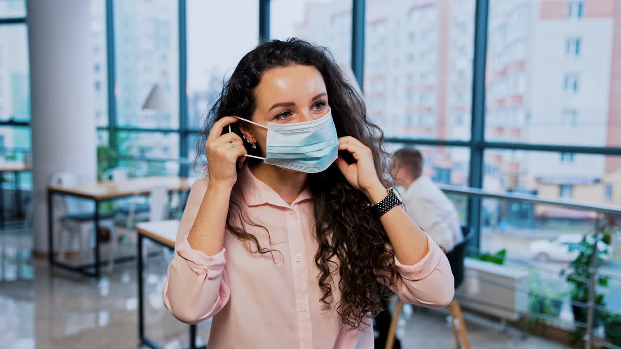 Long-haired lady stand in the office and puts on mask. Office people sitting behind at the table. Panoramic windows at backdrop.