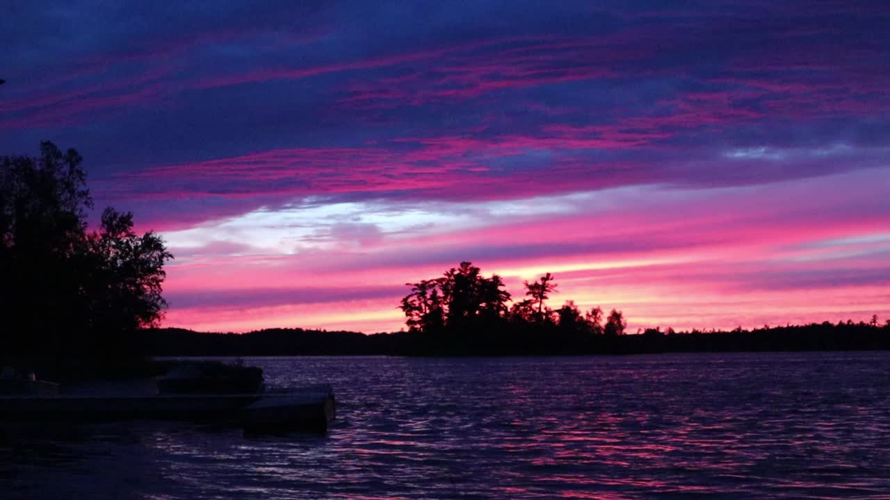 Sunset at Caliper Lake in Northwest Ontario, Canada. A dock extends into the water beneath a colorful sky with red, orange, and blue tones, waves moving across the lake, and a tree silhouette
