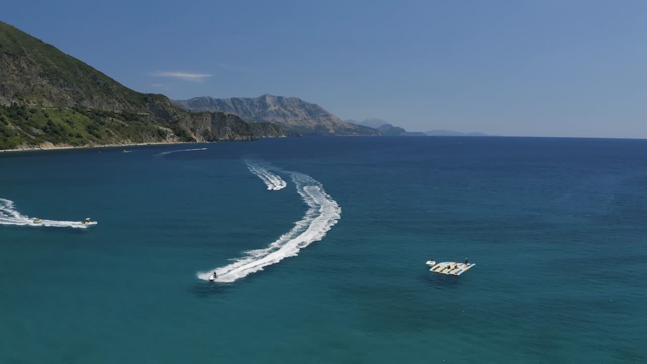 Vista aerea con drone di un motoscafo che si muove attraverso un mare azzurro e limpido, lasciando una scia curva vicino a una costa rocciosa con colline e acqua calma sotto un cielo luminoso diurno.