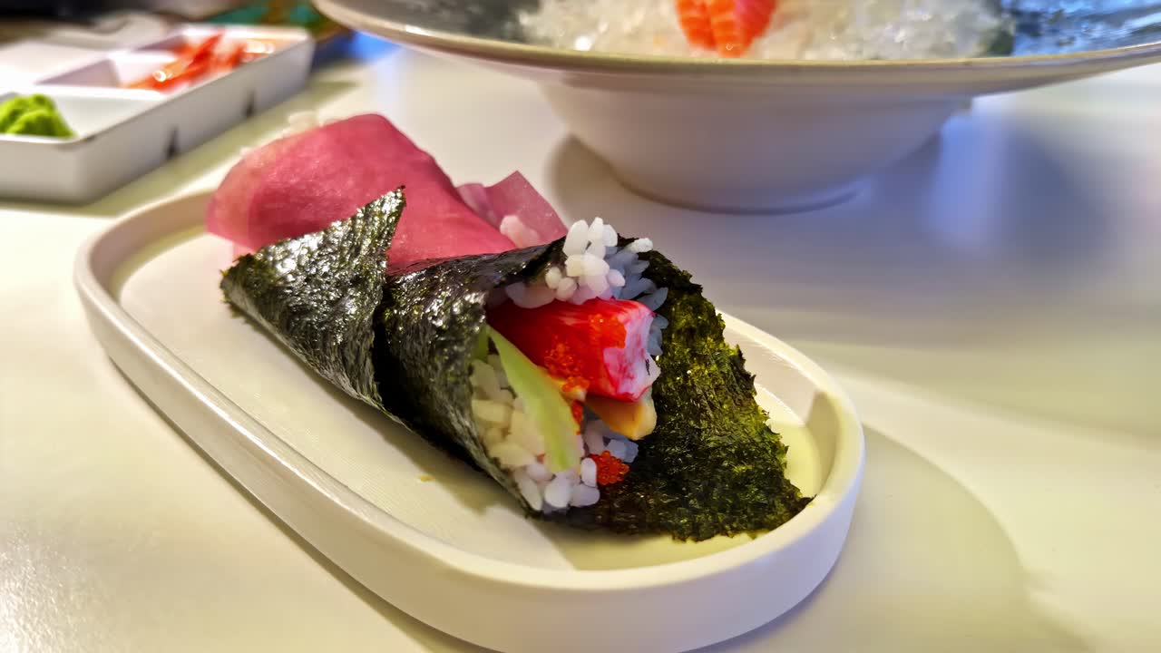 Dekat temaki gulungan tangan yang disajikan dengan elegan bersama acar jahe. Bahan-bahan yang penuh warna dan presentasi yang apik menonjolkan tekstur, gaya, dan estetika bersantap Jepang modern.
