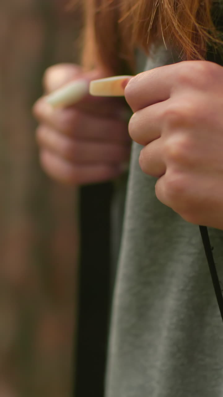 primer plano de una mujer caminando a través de un bosque exuberante, agarrando correas de mochila con uñas largas y bien cuidadosas, el cabello largo fluye sobre sus hombros usando una sudadera gris, contra una vegetación suave y borrosa