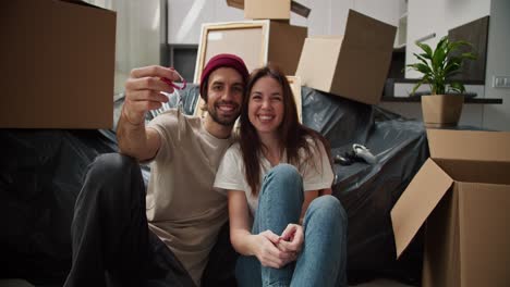 portrait of a happy brunette man with stubble in a red hat and a beige t-shirt who holds in his hands the keys to his new apartment sitting on the floor with his girlfriend near the sofa packed in a black plastic case among a large number of boxes in his new apartment after moving