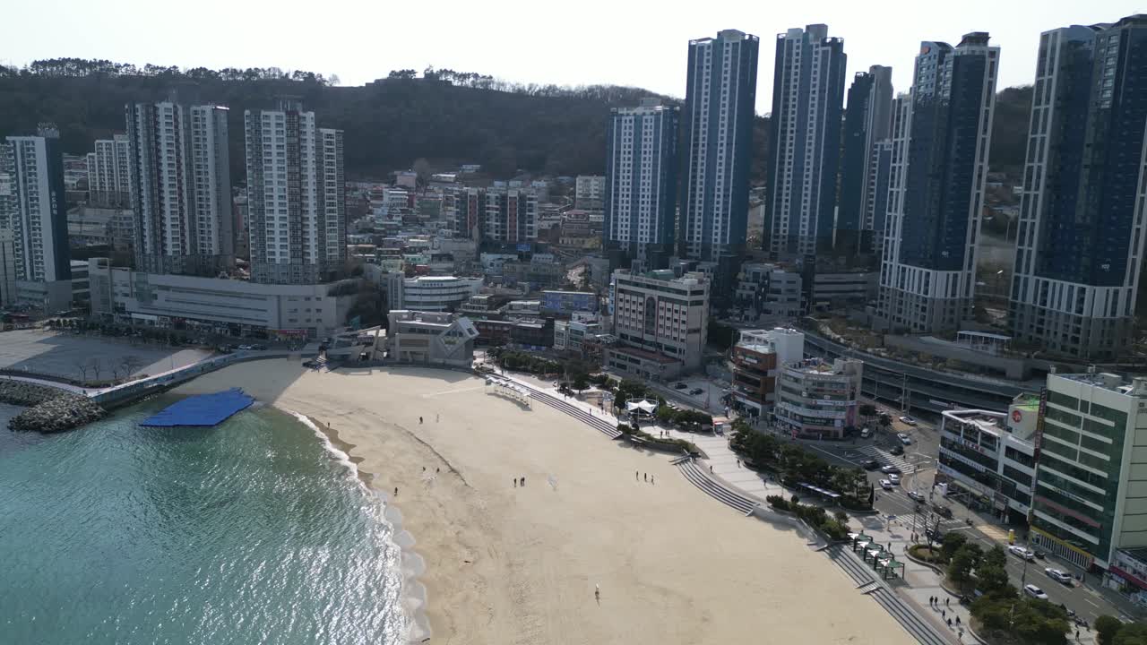 Drone aerial view in South Korea Busan flying over the yellow sand beach with big buildings on the side with people walking on the beach on a sunny day