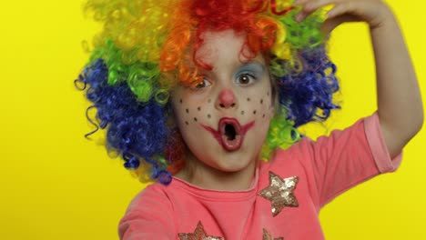 little child girl clown in colorful wig hides behind her hands and shows funny faces. halloween