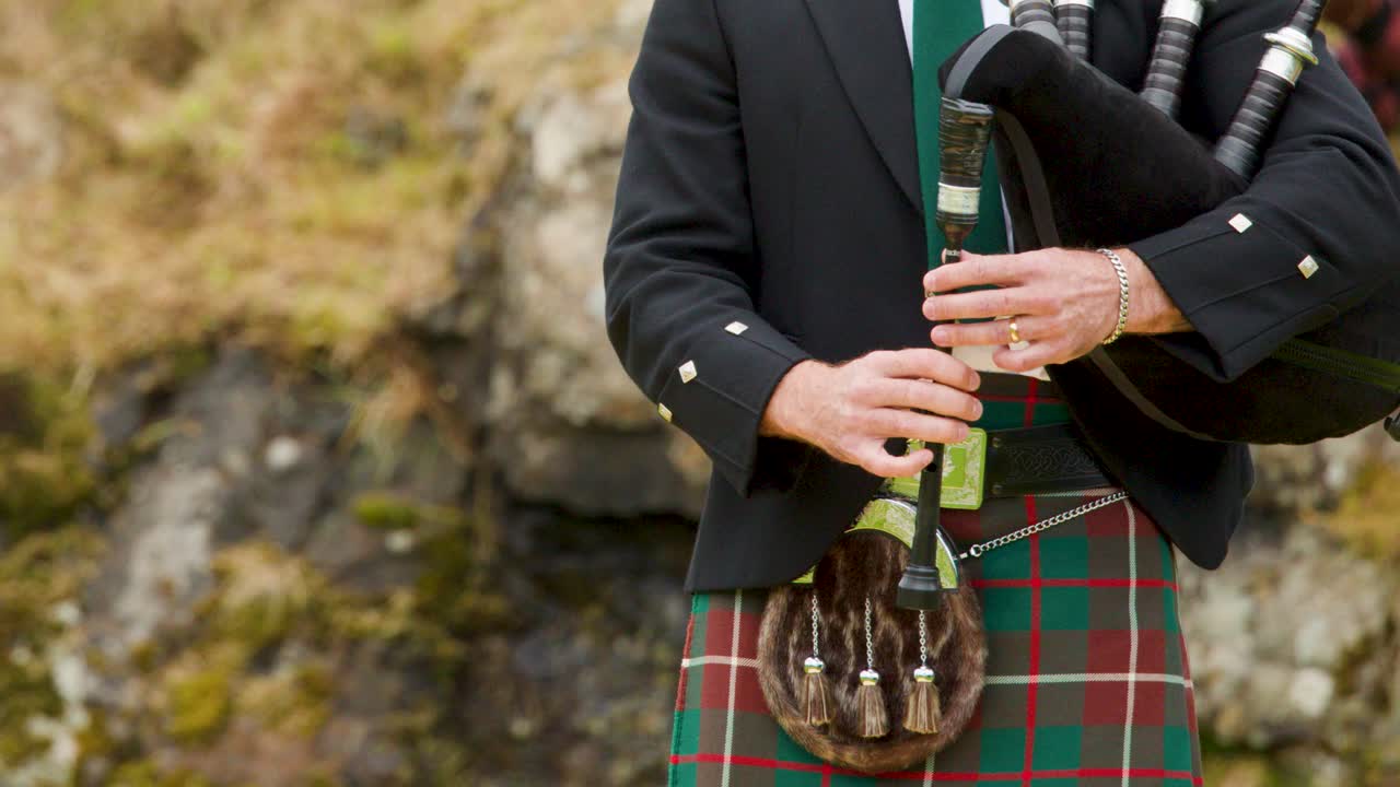 Mann i tartan-kilt spiller sekkepipe utendørs, naturlig dagslys, stødig kamera, bakgrunn med skotsk høylandskap